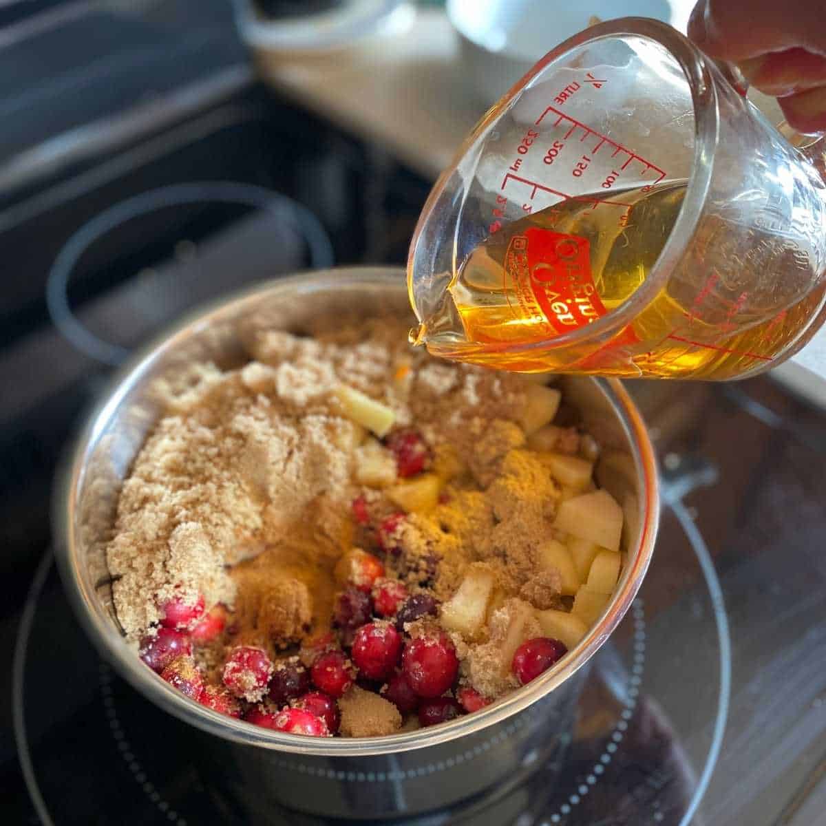 Apple juice being poured into a large saucepan that contains cranberries, apples, brown sugar, cinnamon, and ginger.