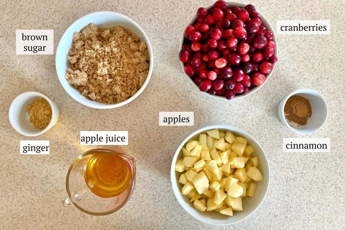 An overhead photo of ingredients needed to make cranberry apple sauce including cranberries, apples, brown sugar, apple juice, ground ginger, and ground cinnamon.