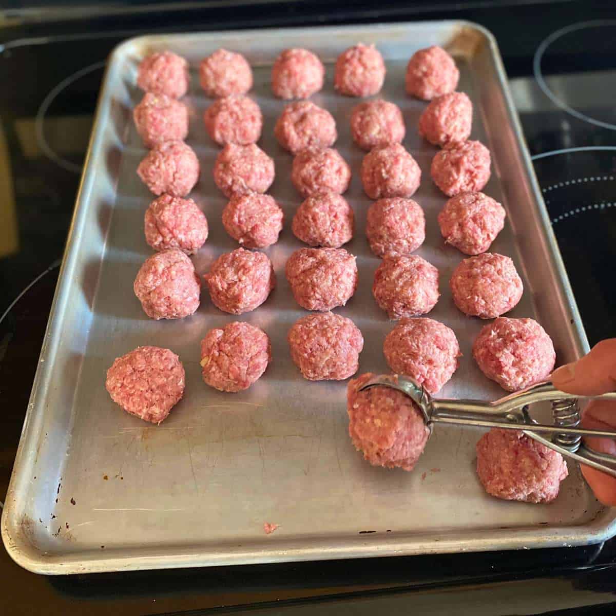 A meatball being formed with a cookie scoop and placed on a baking sheet of meatballs.