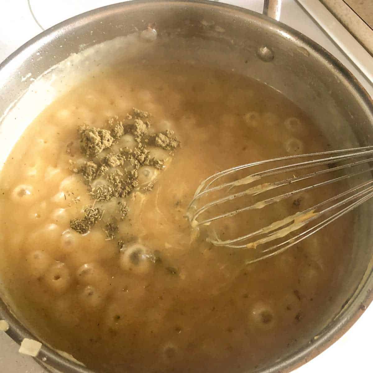 Sauce bubbling in a skillet with sage and thyme being stirred in with a whisk.