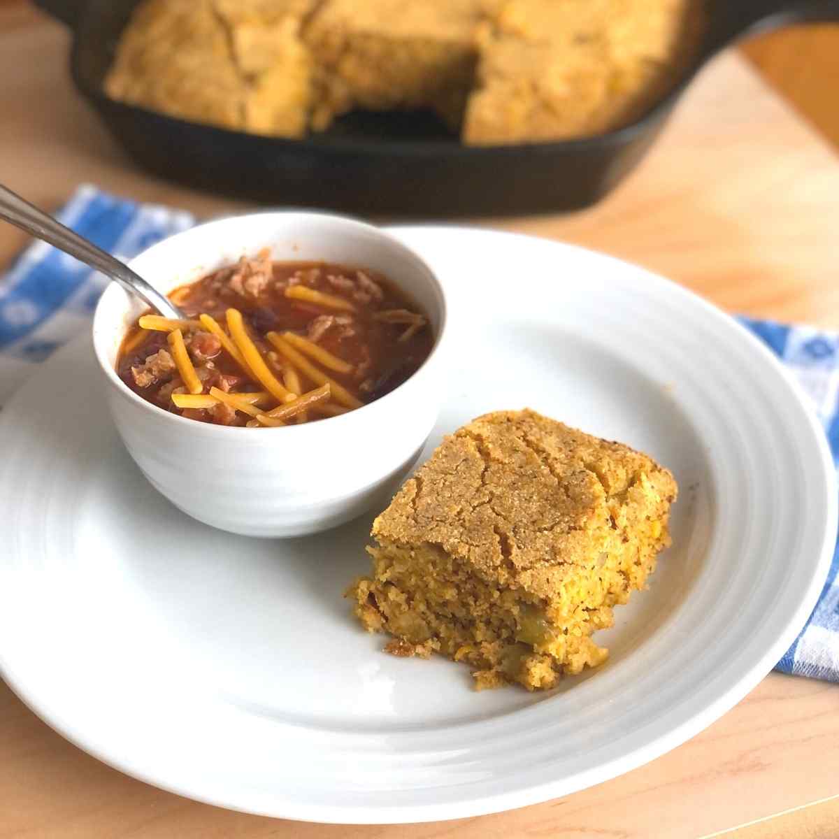 A piece of gluten free cornbread on a plate with a bowl of chili topped with dairy free cheese on the side