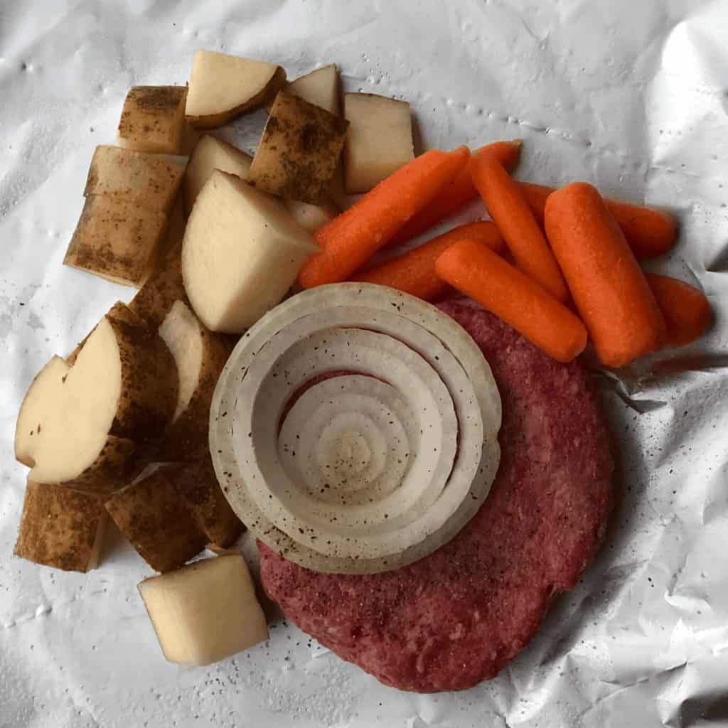 A burger patty, potatoes, baby carrots, and a slice of onion on foil.
