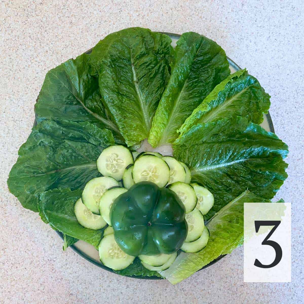 A round plate covered with lettuce, a mound of cucumbers and the end of a bell pepper on top.